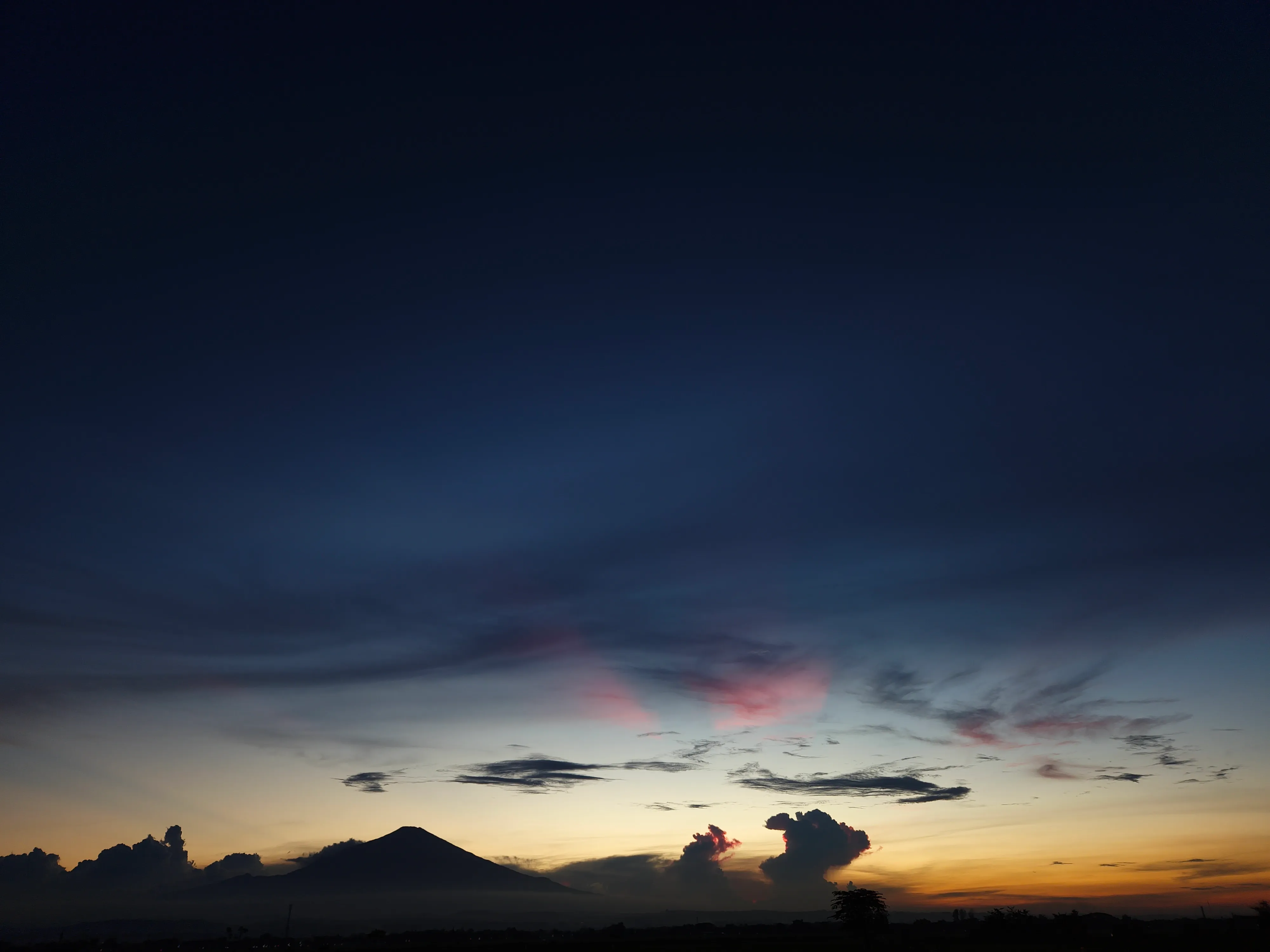 A deep navy blue twilight sky features small pink and red radiating cloud streaks fanning outward across the mid-frame, above a low horizon with a cone-shaped mountain peak silhouette and a dark cumulonimbus cloud mass.
